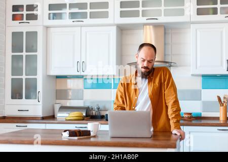 Man use laptop for online work education or shopping. Portrait of bold Bearded Caucasian man Freelancer using his office in kitchen Workspace wooden Stock Photo