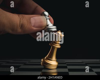 Close-up little silver pawn piece attack the golden queen on chessboard on dark background. Hand moving chess figure on chess game competition. Strate Stock Photo