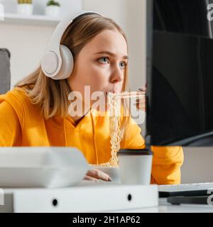 Hungry female gamer eats noodles Chinese dish with chopsticks at home interior using desktop pc computer during streaming video game. Woman teenage Stock Photo