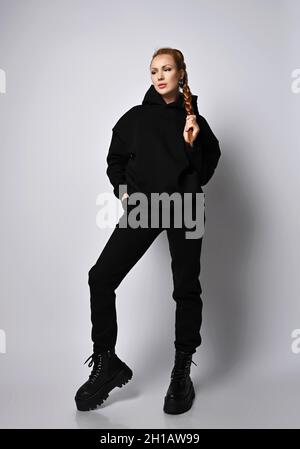 Cool red-haired woman in trendy black sportswear hoodie, pants and massive shoes stands holding her braid with hand Stock Photo