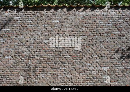 Old grunge brick stone wall made with baked rectangular and square bricks. Roof tiles on top of the wall with leaves behind. Shadows of branches Stock Photo