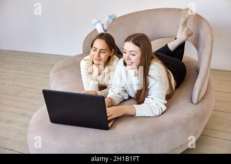 Two happy young sisters lie on sofa with laptop. Stock Photo