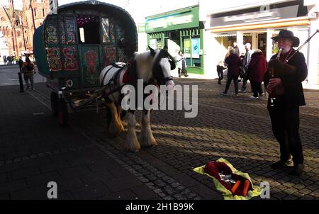 Bow Top Gypsy horse drawn painted traditional Caravan busker in Dumfries, Scotland  playing bagpipes;  with donated cash and carrots.  The design  of the Bow Top wagon traditionally incorporates a lightweight canvas top, on  a wooden frame thought to be based on  older “bender tents” used by the Romanichal gypsies. They were highly decorated inside and out and were  painted green to be less noticeable when parked in  woodland areas or fields. Stock Photo