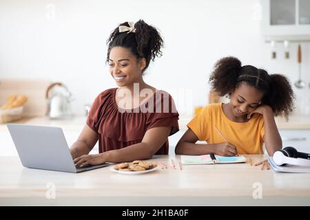 School girl doing homework by her working mother, kitchen interior Stock Photo
