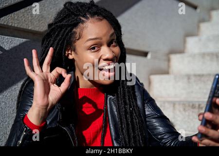 A portrait of a happy African-American woman with dreadlocks in her hair, wearing headphones and listening to music in a park. Stock Photo