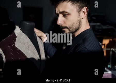 Handsome serious tailor in suit working in atelier with mannequin Stock Photo