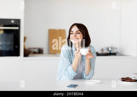 Start of the day. Happy dreamy lady enjoying morning coffee in kitchen interior, holding cup of hot drink and smiling Stock Photo
