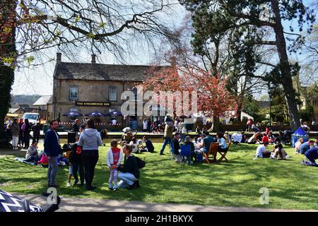 People gather on the central green in Bourton-on-the-Water to enjoy a picnic in the sunshine. The Kingsbridge Inn is in the background. Stock Photo
