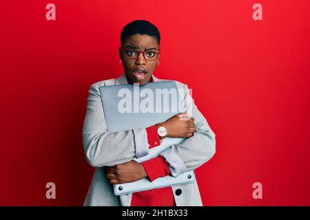 Young african american businesswoman holding laptop in shock face, looking skeptical and sarcastic, surprised with open mouth Stock Photo