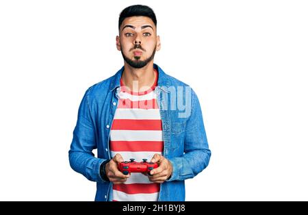 Young hispanic man with beard playing video game holding controller puffing cheeks with funny face. mouth inflated with air, catching air. Stock Photo