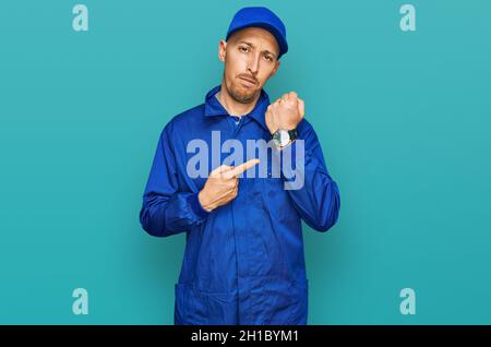 Bald man with beard wearing builder jumpsuit uniform in hurry pointing to watch time, impatience, looking at the camera with relaxed expression Stock Photo