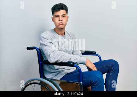Young hispanic man sitting on wheelchair puffing cheeks with funny face. mouth inflated with air, crazy expression. Stock Photo