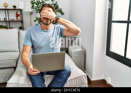 Handsome middle age man using computer laptop on the sofa smiling and laughing with hand on face covering eyes for surprise. blind concept. Stock Photo