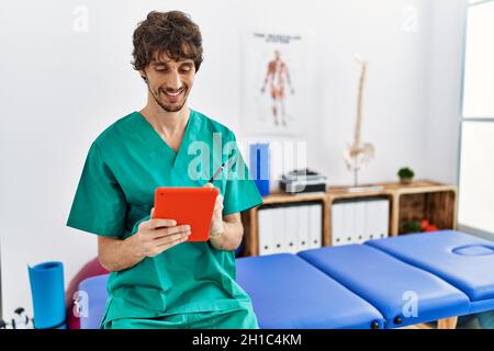 Young hispanic man wearing physiotherapist uniform using touchpad at physiotherapist clinic Stock Photo