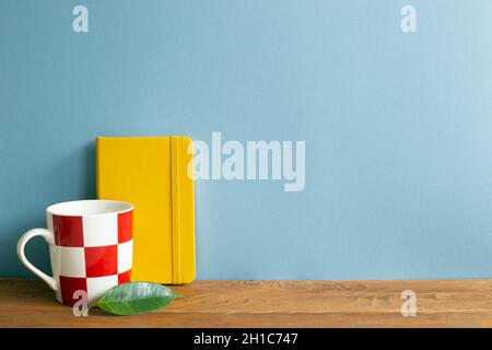 Yellow diary note book with mug cup on wooden desk. blue wall background Stock Photo