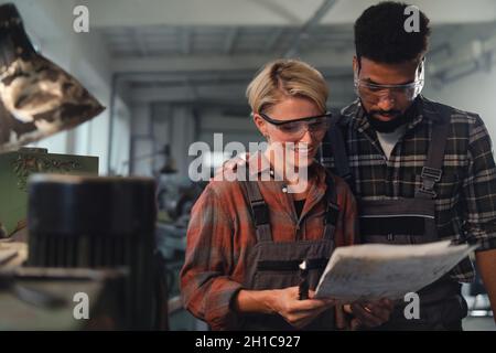 Portrait of young biracial industrial colleagues working indoors in metal workshop, smiling. Stock Photo