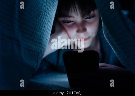 Teen girl using smartphone, hiding under blanket at nigh, social networks cocnept. Stock Photo