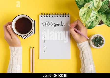 Desktop blank notepad and woman hands writing 2022 plans and goals text . Flat lay of yellow working table background with cup of tea and plants. Top Stock Photo