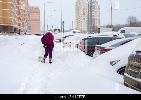 Moscow, Russia - February 13, 2021: Parking on the street covered with snow. The woman walks the dog among the huge snowdrifts Stock Photo