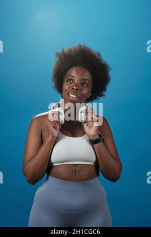 Young plus size female in activewear touching headphones while standing on blue background