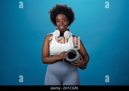 Young pretty African sportswoman with rolled mat standing against blue background