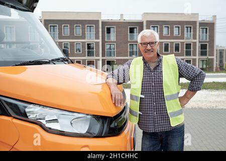 Happy mature man in workwear standing by front part of bus against modern architecture Stock Photo