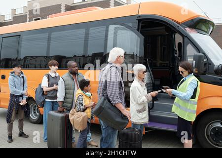 Young bus conductor checking tickets by open door of vehicle with line if passengers in front Stock Photo