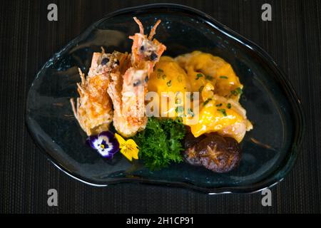 Two shrimps in batter with sauce and mushrooms on the blue designed plate. Deep fried prawns decorated with greenery and flower. Isolated on black Stock Photo