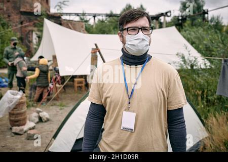 Young volunteer in protective mask standing in refugee camp against group of migrants Stock Photo