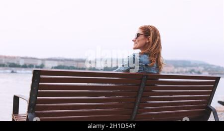 Happy young redhead woman relaxing on bench at riverside in city Stock Photo