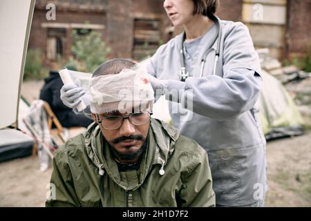 Young doctor in uniform putting bandage around wounded head of refugee man Stock Photo