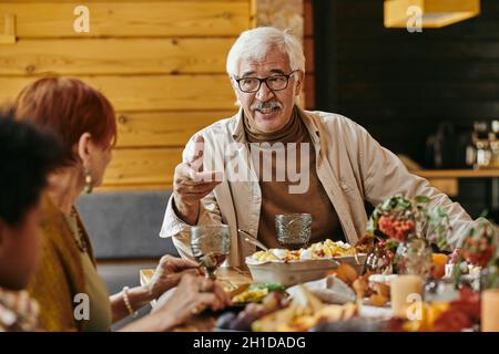 Senior man in eyeglasses talking to the members of his family while sitting at dining table at home Stock Photo