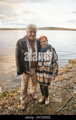 Portrait of happy senior couple embracing and smiling at camera while standing near the lake outdoors Stock Photo