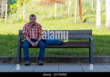 Gipuzkoa, SPAIN-MARCH 22, 2018 : Sad and tired man sitting on wooden bench in park. Old man wear plaid shirt and jeans. Caucasian senior man sitting a Stock Photo