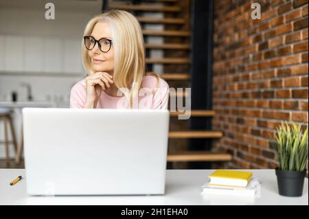 Thoughtful elegant middle-aged businesswoman wearing stylish glasses using with a laptop indoor, sits at the desk and looks away dreamly. Charming blonde woman working in the loft office, taking break Stock Photo