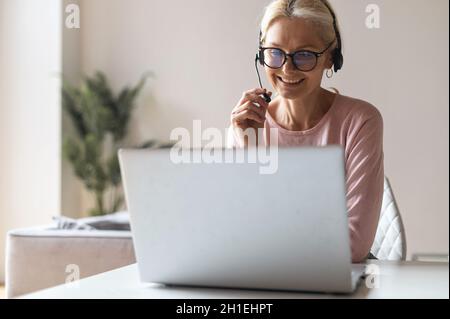 Smiling female call center employee using a headset and laptop for online communication with customers, a woman talking into microphone and typing on the keyboard, checks and marks Stock Photo