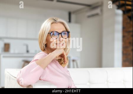 Elegant charming middle-aged woman with blond hair sitting on the comfortable couch and looks at the camera, smiling, put her chin on hands. Wellbeing concept Stock Photo