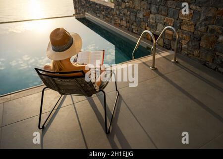 relaxing vacation - woman sitting in chair and reading a book on terrace near swimming pool at luxury villa Stock Photo