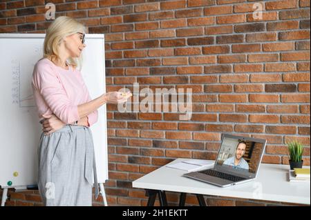 A friendly middle-aged female office worker wearing glasses in the office, looking at the laptop screen, talking with participant of online conference, speacking to male student, coworker Stock Photo