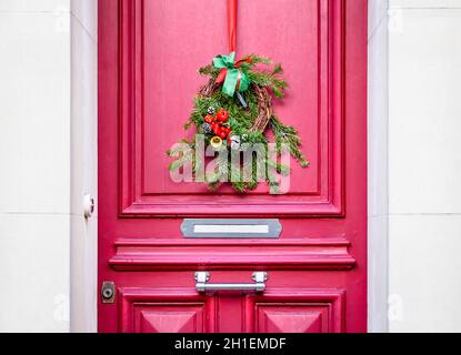 Front view of a Christmas crown made of fir branches, pine cones and ribbons, hanging on the red front door with moldings of a townhouse. Stock Photo