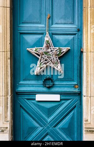 Front view of a star-shape Christmas decoration made of branches and pine cones, hanging on the blue front door with moldings of an old townhouse. Stock Photo