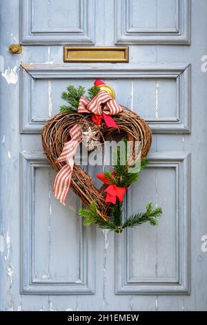 Front view of a heart-shape Christmas decoration made of branches and ribbons, hanging on a worn blue front door with moldings. Stock Photo