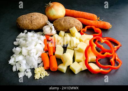 Prepped Raw Vegetables on a Dark Background: Peeled, sliced, and chopped onion, potatoes, carrots, red bell peppers, and garlic Stock Photo