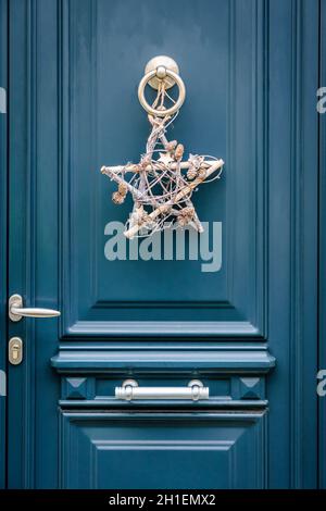 Front view of a star-shape Christmas decoration made of branches and pine cones, hanging on a blue front door with moldings. Stock Photo