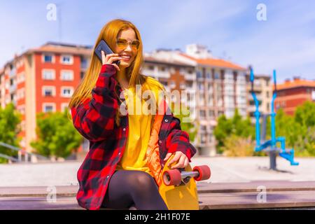 Stylish redhead Hispanic skater lady talking on her phone enjoying the sunny day in an urban park Stock Photo