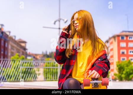 Stylish redhead Hispanic lady talking on her phone in an urban park holding her skateboard Stock Photo