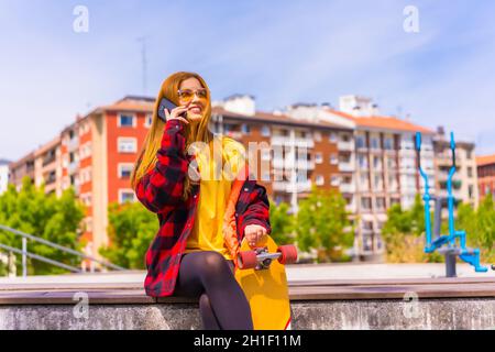 Stylish redhead Hispanic lady talking on her phone in an urban park holding her skateboard Stock Photo