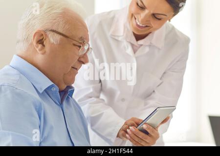Smiling nurse teaching senior man to use healthcare app or showing photo of his grandchildren Stock Photo