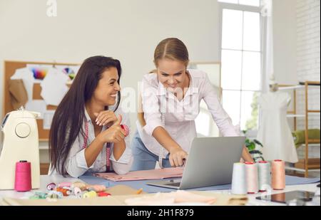 Two young female seamstresses in sewing studio check and record customer orders using laptop. Stock Photo