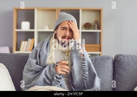 Sick man with a glass of water in his hand suffers from a great headache sitting on the couch. Stock Photo